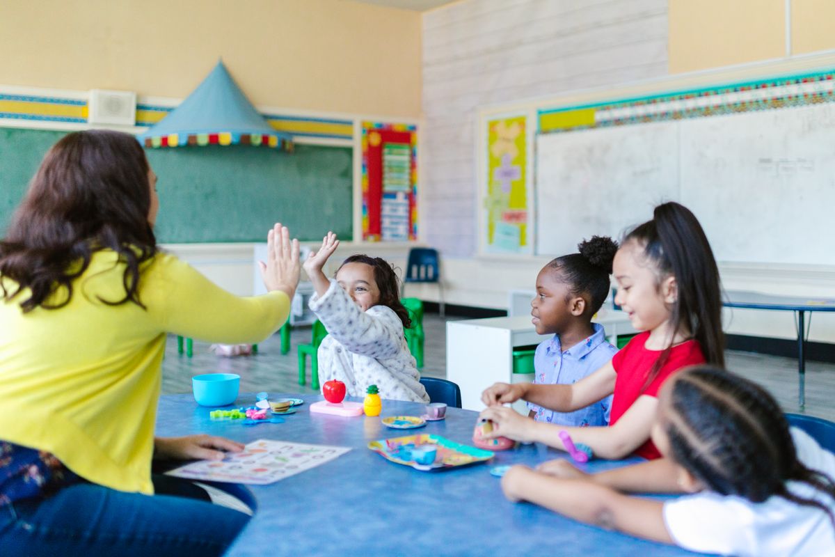 A teacher high-fives a child while other children play at a classroom table